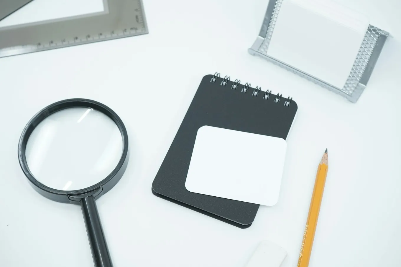 Photo: a magnifying glass, a small black notebook, a pencil and a ruler laid out on a white desk.